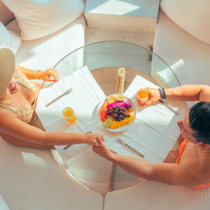 A couple in swimwear, reflecting the allure of Soneva Exclusive Offers, sits at a round glass table on a boat, holding hands. The table is set with a fruit platter and drinks. The woman wears a sunhat, and the scene is bright and sunny, suggesting a relaxed, vacation-like atmosphere.