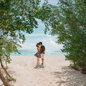 A couple stands embracing under a leafy archway on a sandy beach with the sparkling ocean and blue sky in the background. The man is dressed in a navy shirt and white shorts, while the woman wears a floral dress. Sunlight filters through the trees, creating an idyllic setting reminiscent of Soneva Exclusive Offers.