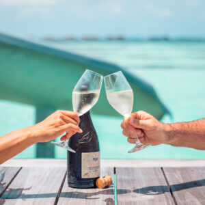 Two hands clink champagne glasses over a wooden table with a bottle of champagne and a cork on it. Clear blue water and a green slide are visible in the background, suggesting an outdoor, waterfront setting ideal for enjoying Soneva&#039;s exclusive offers.