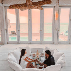 A couple sits across from each other at a round glass table, holding hands in a white, cushion-filled seating area. Large windows offer a view of the ocean and a pink sunset. Above them hangs a decorative fixture made of intertwined branches, adding to the romance reminiscent of Soneva Exclusive Offers.