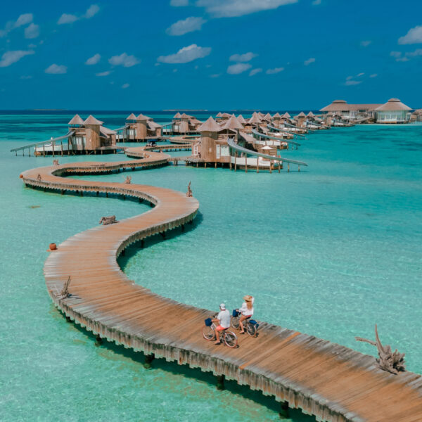 A scenic view of a long, curved wooden boardwalk stretching over turquoise waters, leading to overwater bungalows. People are riding bicycles and walking along the path under a clear blue sky.