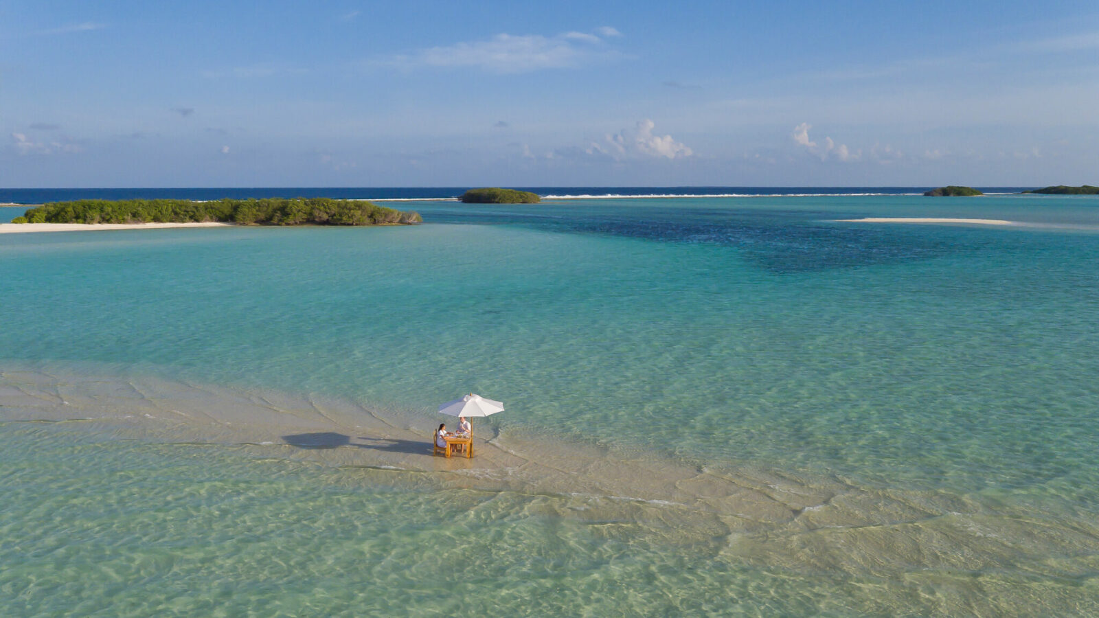 A tranquil beach scene with clear, shallow turquoise water and a sandy area in the foreground. A small wooden table and chair with a white parasol are set up on the sand. Lush green islands and a calm blue sky with scattered clouds are visible in the background—perfect for a Soneva Maldives holiday.