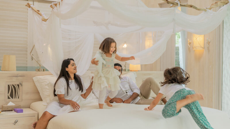 In a bedroom bathed in natural light and decorated with soft, neutral tones, a joyful family enjoys precious moments together. As two children gleefully jump on a large, canopied bed, their parents watch with smiles. Discover exclusive offers at Soneva Fushi for your own family retreat.