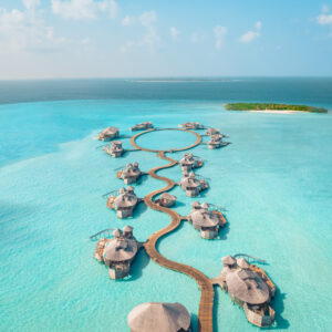 Aerial view of luxurious overwater villas at the Maldives luxury resort Soneva, connected by winding wooden walkways in a turquoise sea. The circular arrangement of some villas stands out, with an island of lush greenery visible in the distance under a clear, blue sky.