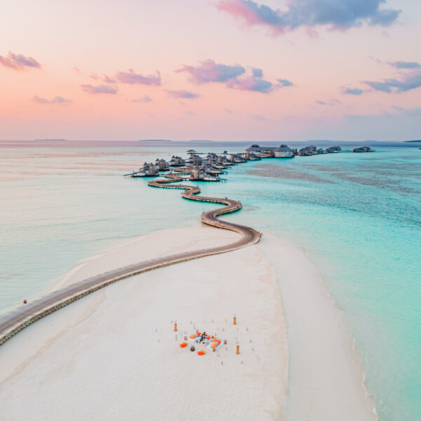 Aerial view of a serene beach at sunset, featuring a winding walkway leading to overwater bungalows. The calm turquoise ocean surrounds the sandy shore, where a small seating area, set with pillows and low tables, is arranged for a relaxing Soneva Maldives holiday experience.