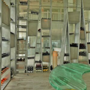 Modern wine cellar with metal shelving units holding a variety of wine bottles. A large, green-tinted, curved glass table is placed in the foreground. The room is well-lit, and the shelves are artistically arranged in an abstract display reminiscent of a Maldives luxury resort.