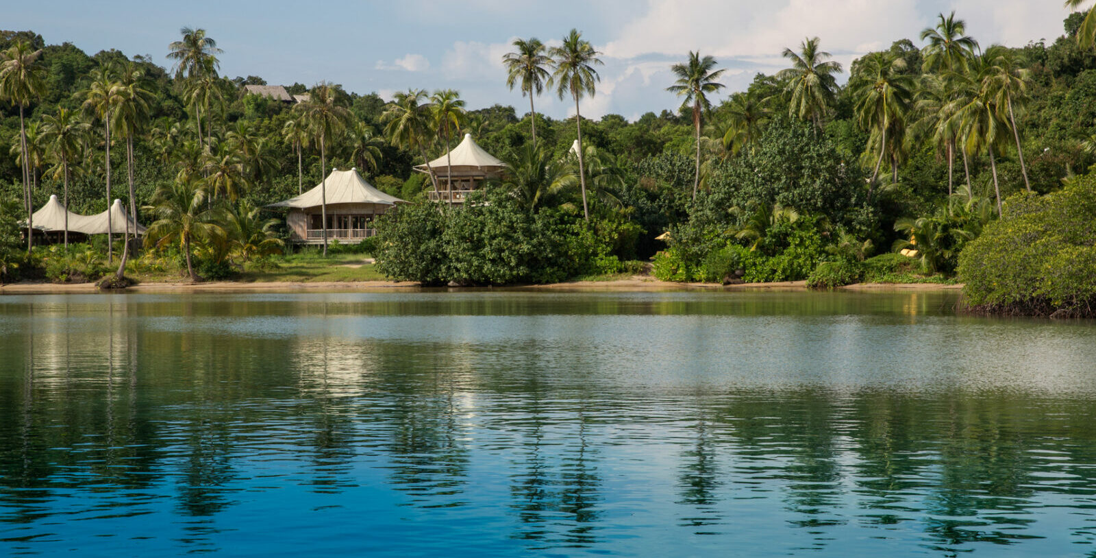 A serene tropical scene featuring a tranquil body of water in the foreground with several luxury tents from Soneva Kiri Resort and lush greenery in the background. Tall palm trees are scattered throughout the landscape under a partly cloudy sky.