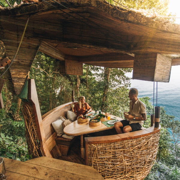 Two people sit in a unique, nest-like dining pod suspended in a tree. They enjoy a meal surrounded by lush greenery with a view of the ocean in the background. The pod is made of woven materials, creating a cozy, rustic atmosphere.