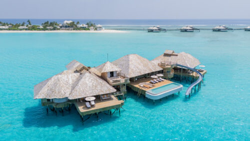 Overwater bungalows with thatched roofs in a tropical setting, surrounded by clear turquoise water. The main Crusoe Reserve bungalow features a pool and slide. A distant island with more bungalows on stilts is visible on the horizon, offering a perfect escape to paradise.