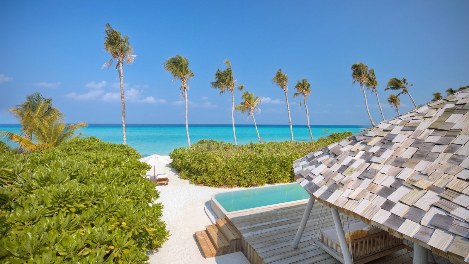 A serene beach scene with clear blue skies, calm turquoise ocean water, and tall palm trees swaying in the breeze. In the foreground, a wooden deck with a thatched-roof structure next to a small pool is bordered by lush green bushes, epitomizing the luxury of Soneva Exclusive Offers.