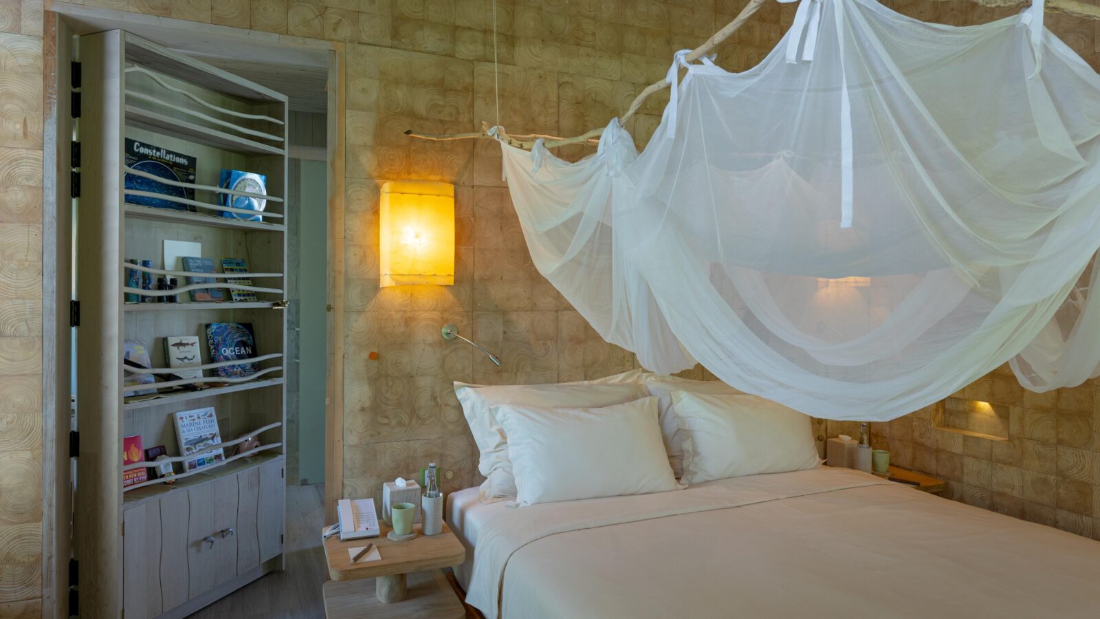 A cozy bedroom with a canopy bed draped in white fabric. The room features light wooden walls and fixtures, including a bedside table with a lamp and essential items. An open door reveals shelves stocked with books and decorative items. Warm lighting adds a serene ambiance, perfect for enjoying Soneva Exclusive Offers.