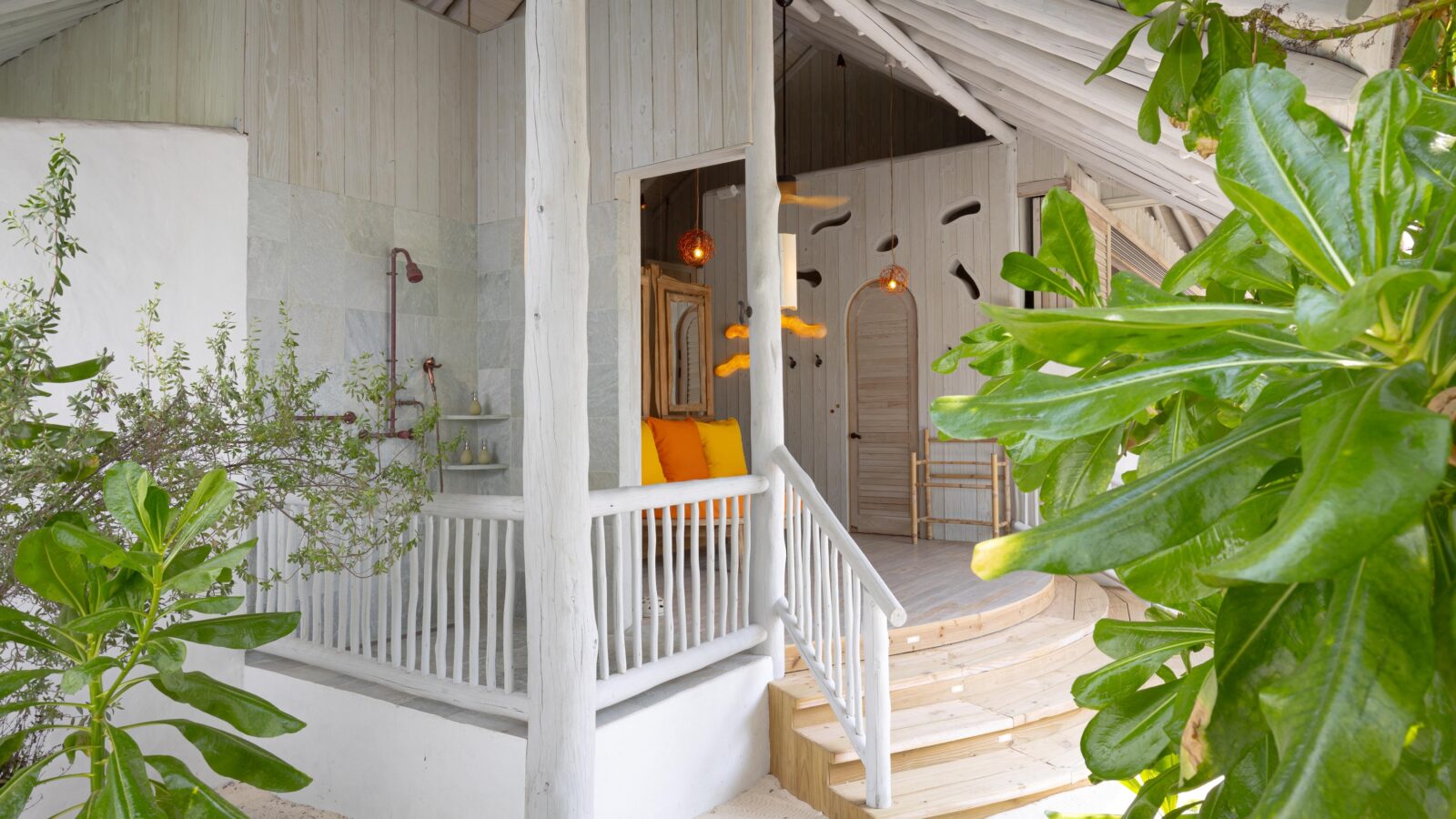 A serene outdoor porch with white wooden railings and steps, adorned with lush green plants. Inside, the space features an inviting seating area with a bright orange cushion, hanging light fixtures, and decorative wall accents in a light, airy setting—perfect for enjoying Soneva Exclusive Offers.