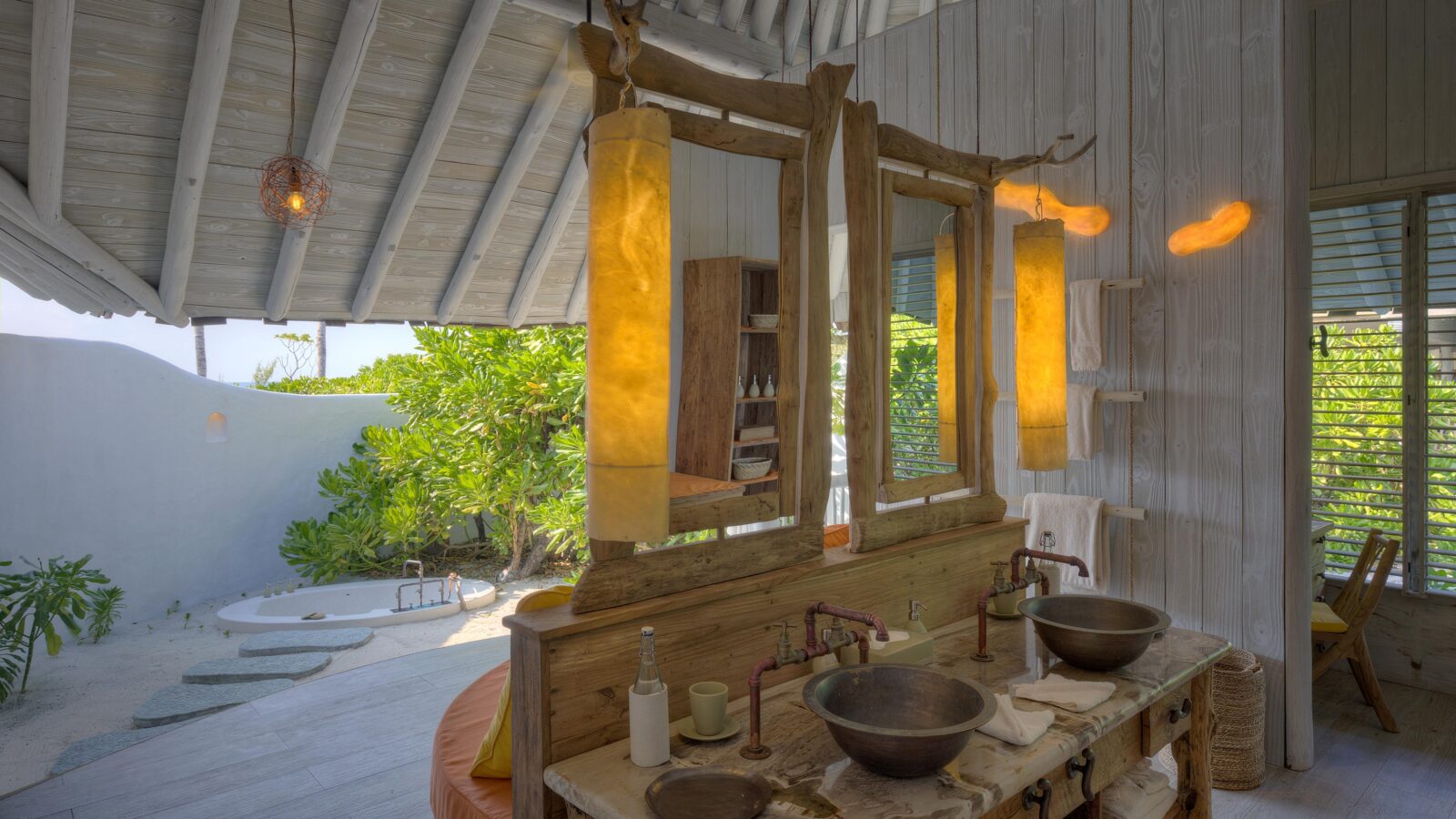 A tropical outdoor bathroom with a rustic wooden vanity featuring two vessel sinks and copper faucets. The area is adorned with warm, ambient lighting, lush greenery, a cozy bathtub, and stepping stones leading to the tub. The space is open and airy with a thatched roof—a true Soneva Exclusive Offers experience.