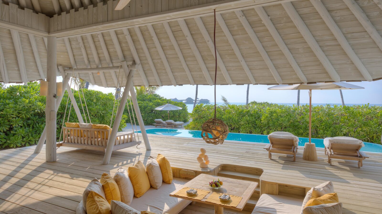 A luxurious, open-air beachside cabana with white wooden beams, a swing, cushioned seating area with yellow pillows, and a dining table awaits you. Beyond the cabana lies a pool surrounded by lush greenery and an ocean view with lounge chairs under umbrellas—experience it all with Soneva Exclusive Offers.