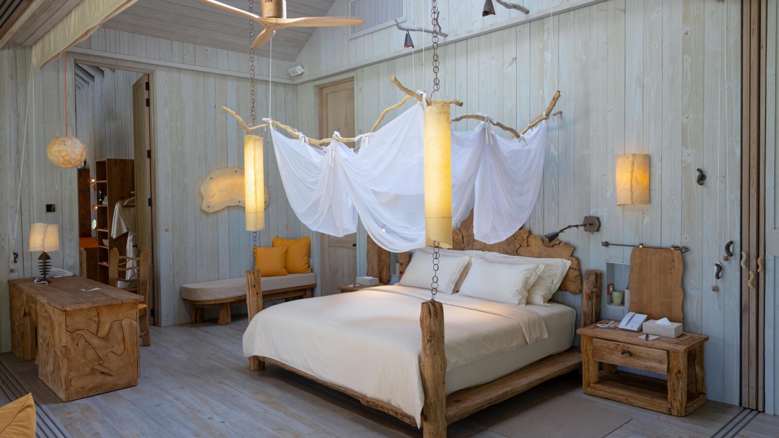 A serene bedroom with light wood decor features a rustic wooden bed draped with flowing white fabric, illuminated by hanging lamps. The room has a natural theme with wooden benches, a ceiling fan, and a minimalist, cozy vibe. Discover unparalleled tranquility with Soneva Exclusive Offers in this peaceful retreat.