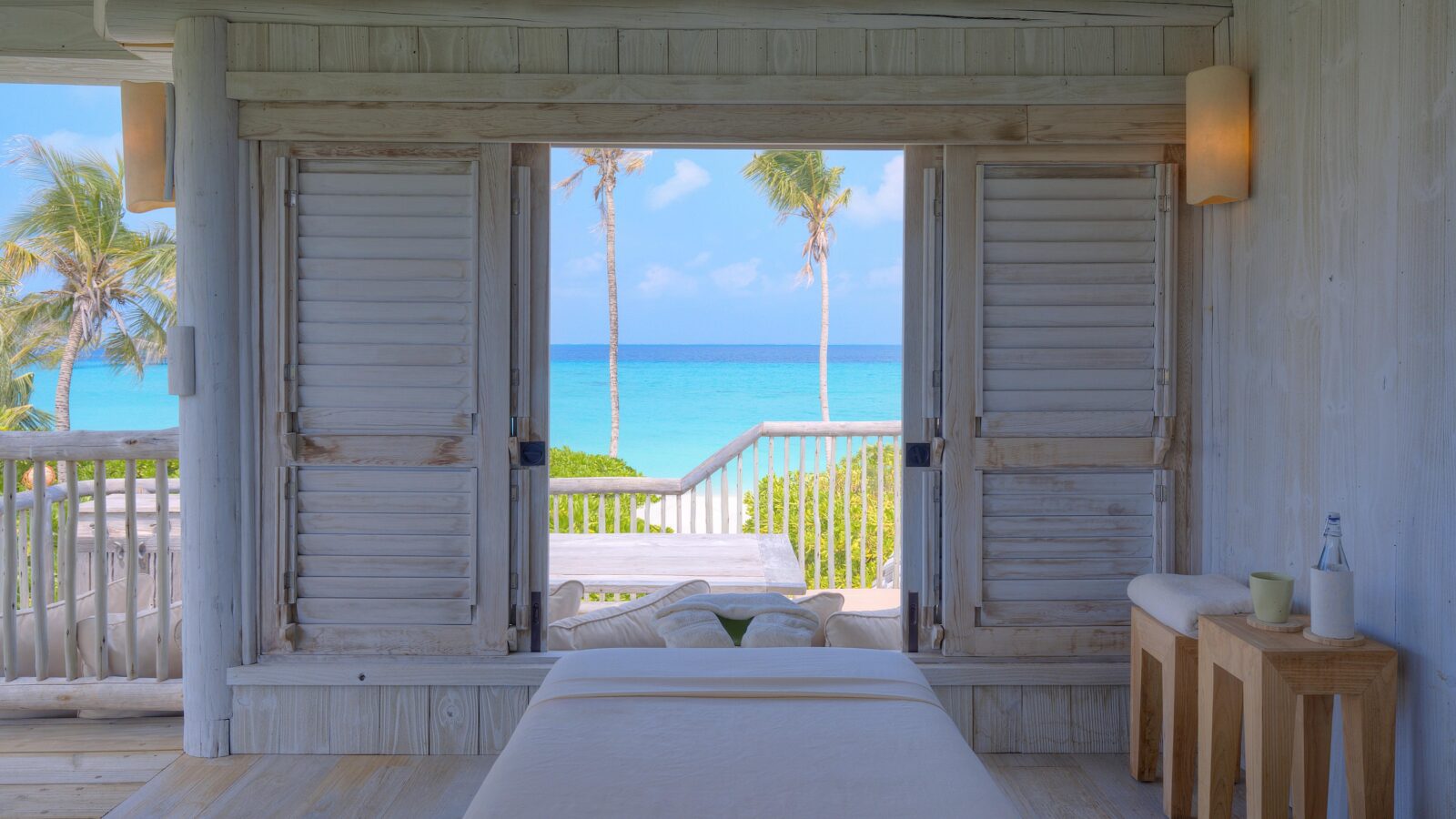 A serene spa room with light wooden decor opens to a stunning ocean view, featuring closed wooden shutters, a massage table with pillows, and two stools with a bottle of water. Set within lush palm trees leading to the bright blue sea, it’s an idyllic retreat perfect for Soneva Exclusive Offers.