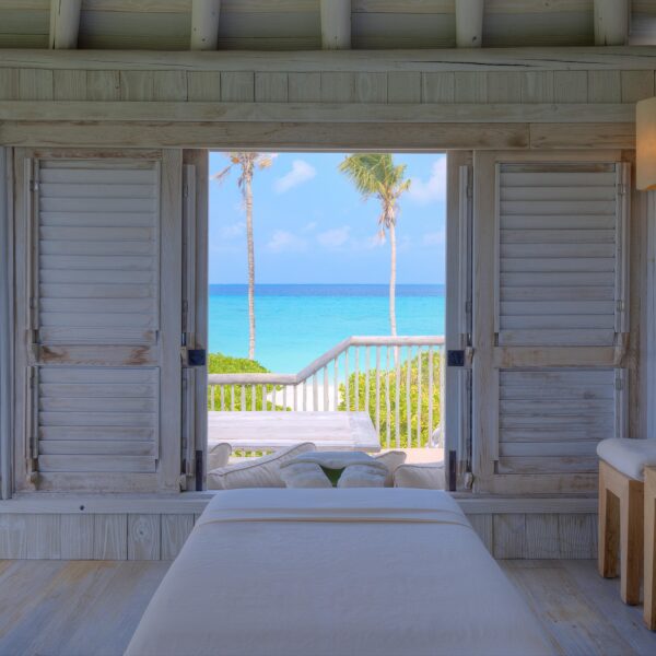 A serene beachside room with wooden shutters open to a view of a turquoise ocean and palm trees. The room features a massage table covered in a white sheet, two wooden stools, and a bottle of water on one stool. Embrace the tranquil and inviting atmosphere with Soneva Exclusive Offers.