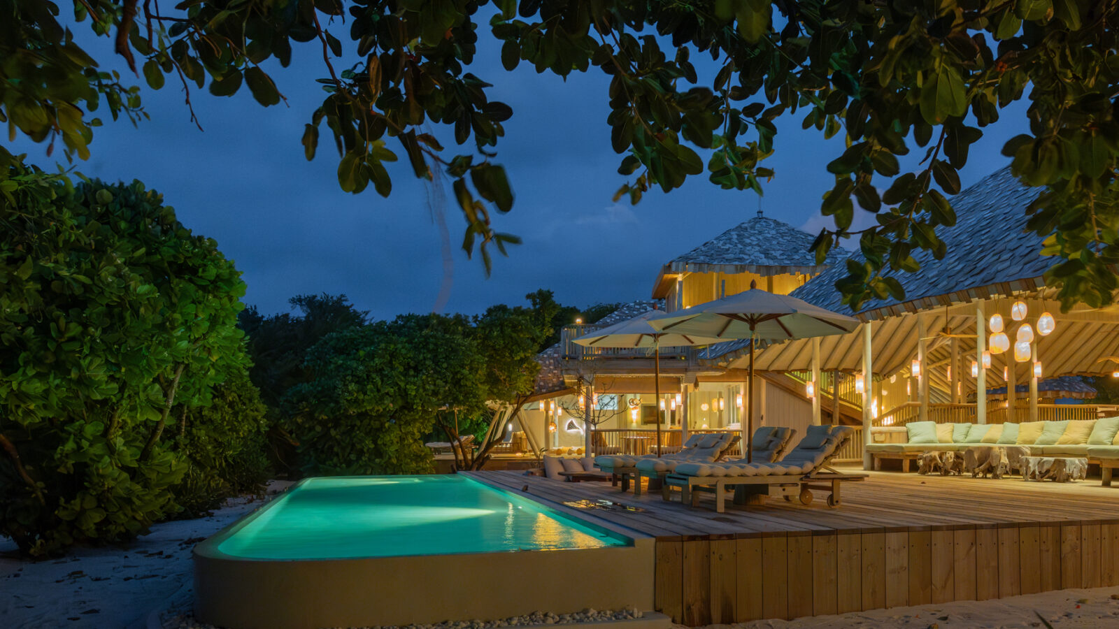 A serene beachfront scene at twilight in a Beach Reserve, featuring a glowing infinity pool and a wooden deck with lounge chairs and umbrellas. A stylish open-air structure with warm lighting is surrounded by lush greenery, as the sky deepens into a rich blue.