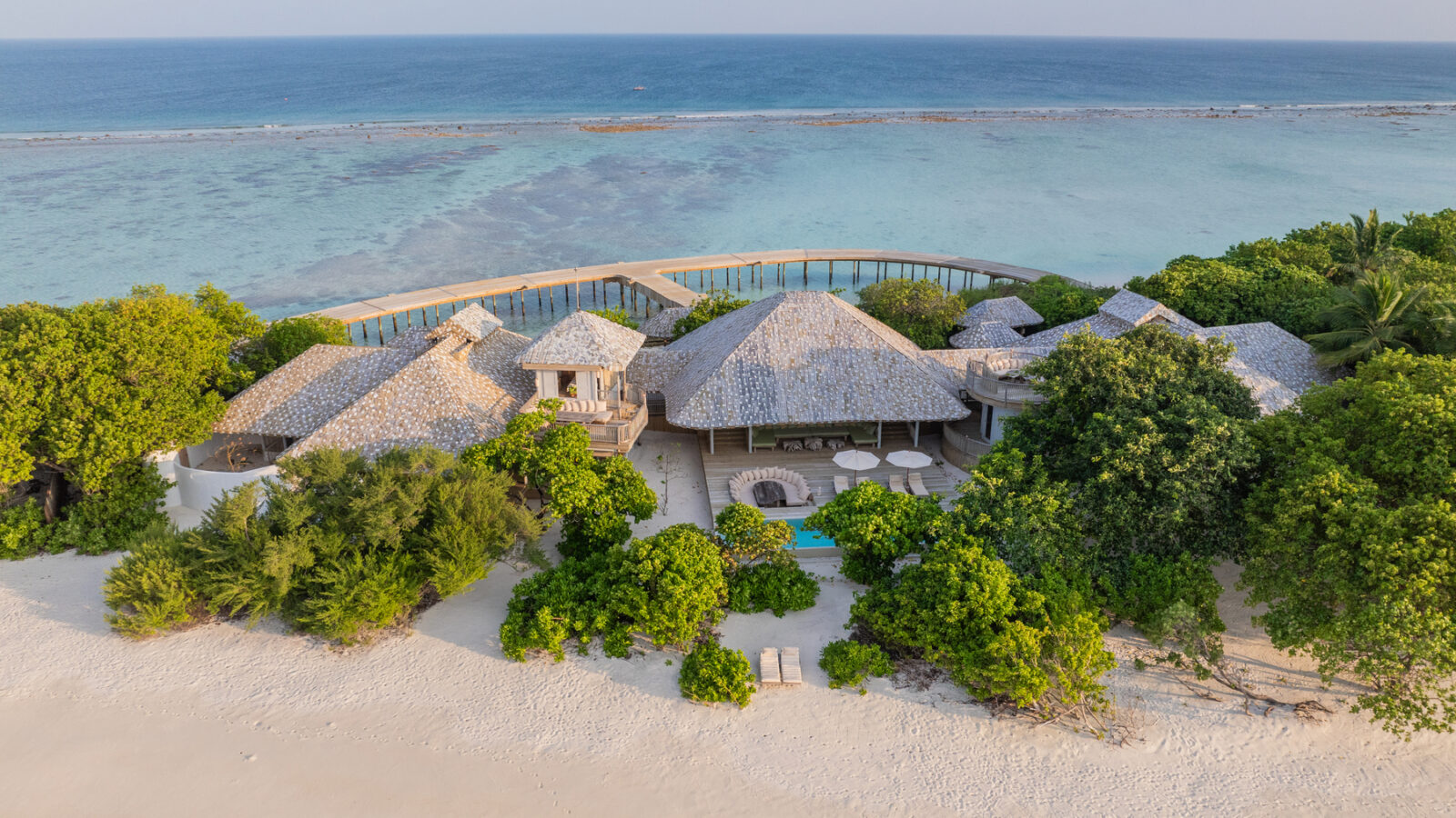 Aerial view of a tropical beach reserve featuring thatched-roof villas nestled in lush greenery. The sandy shore leads to clear blue ocean waters beneath a partly cloudy sky, with a boardwalk extending gracefully over the water.