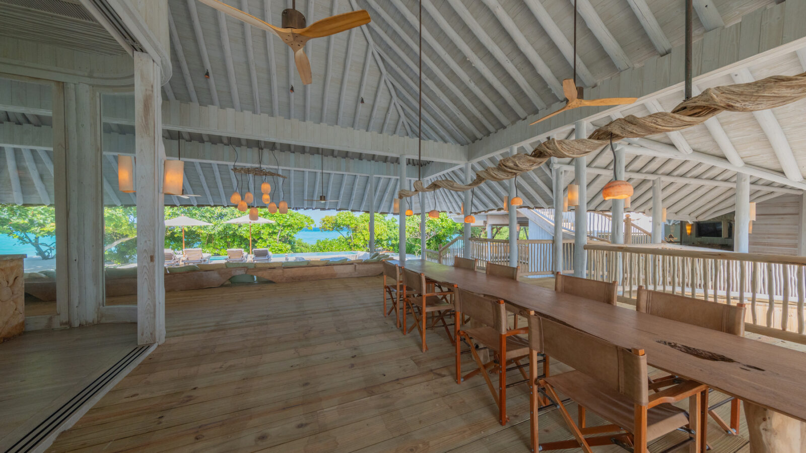 Open-air wooden dining area with sloped ceiling, fans, a long table, and chairs. Large windows unveil lush greenery and the ocean within the Beach Reserve. Pendant lights hang from the ceiling, enhancing the bright and airy atmosphere.