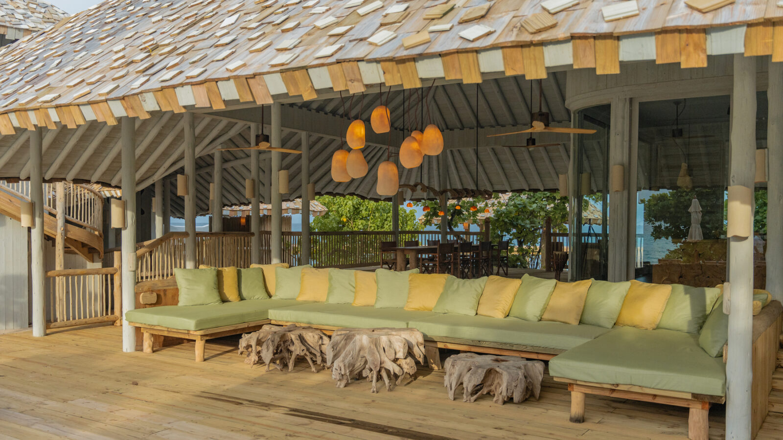 A spacious outdoor seating area at the Beach Reserve features a long, L-shaped sofa with green and yellow cushions under a thatched roof. Unique wooden tables with organic shapes sit in front. Natural light illuminates the cozy, tropical setting.