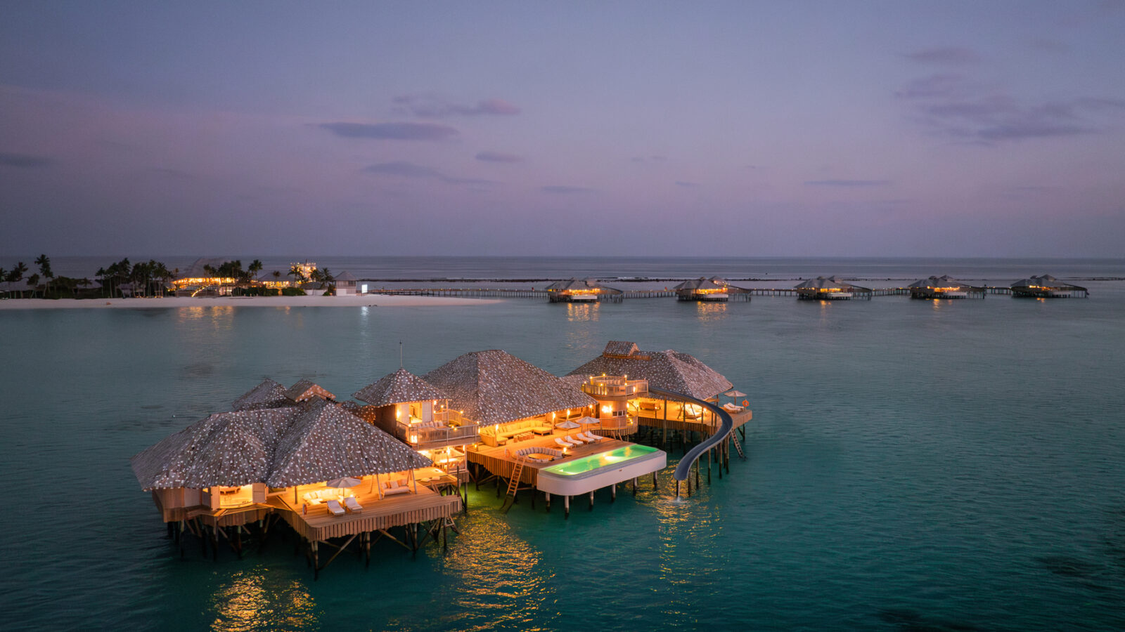 Aerial view of luxurious overwater villas with thatched roofs in a tranquil ocean setting at dusk, reminiscent of a Crusoe Reserve. Warm lights illuminate the villas, reflecting on the calm, turquoise water. A sandy coastline with a few palm trees is visible in the background.