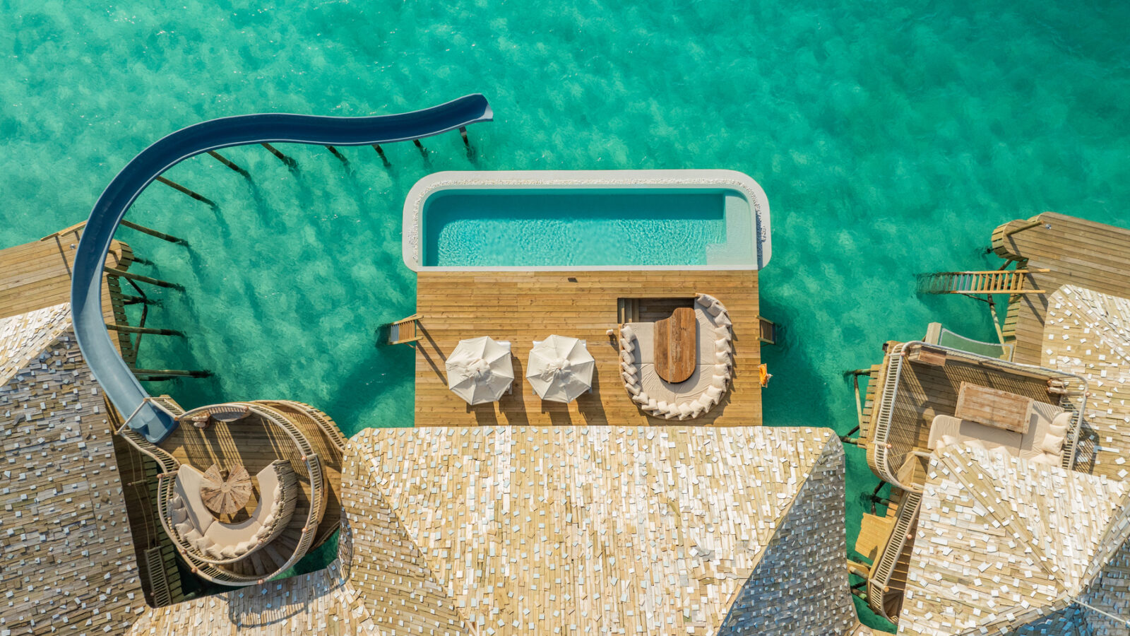 Aerial view of a luxury overwater Crusoe Reserve bungalow with a private pool in clear turquoise water. The bungalow features a waterslide, wooden deck, sun loungers, and shaded seating area, offering a picturesque tropical retreat.