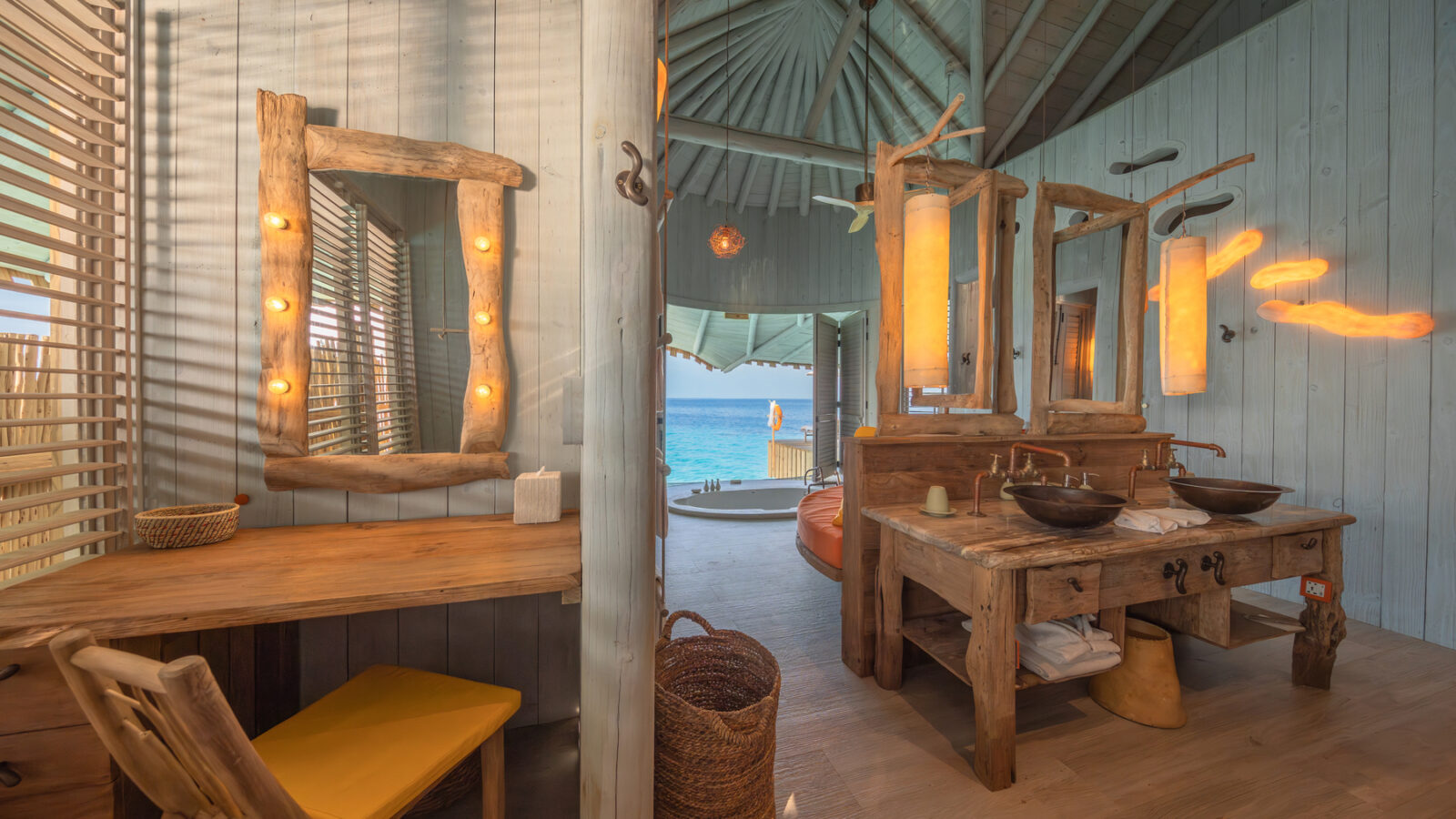 Nestled in the tranquil ambiance of Crusoe Reserve, this cozy rustic beachside room features wooden furniture. A desk with a mirror and lightbulbs, a wicker basket, and candles complement the decor. Large open windows frame an ocean view, while light wood walls enhance the natural atmosphere.