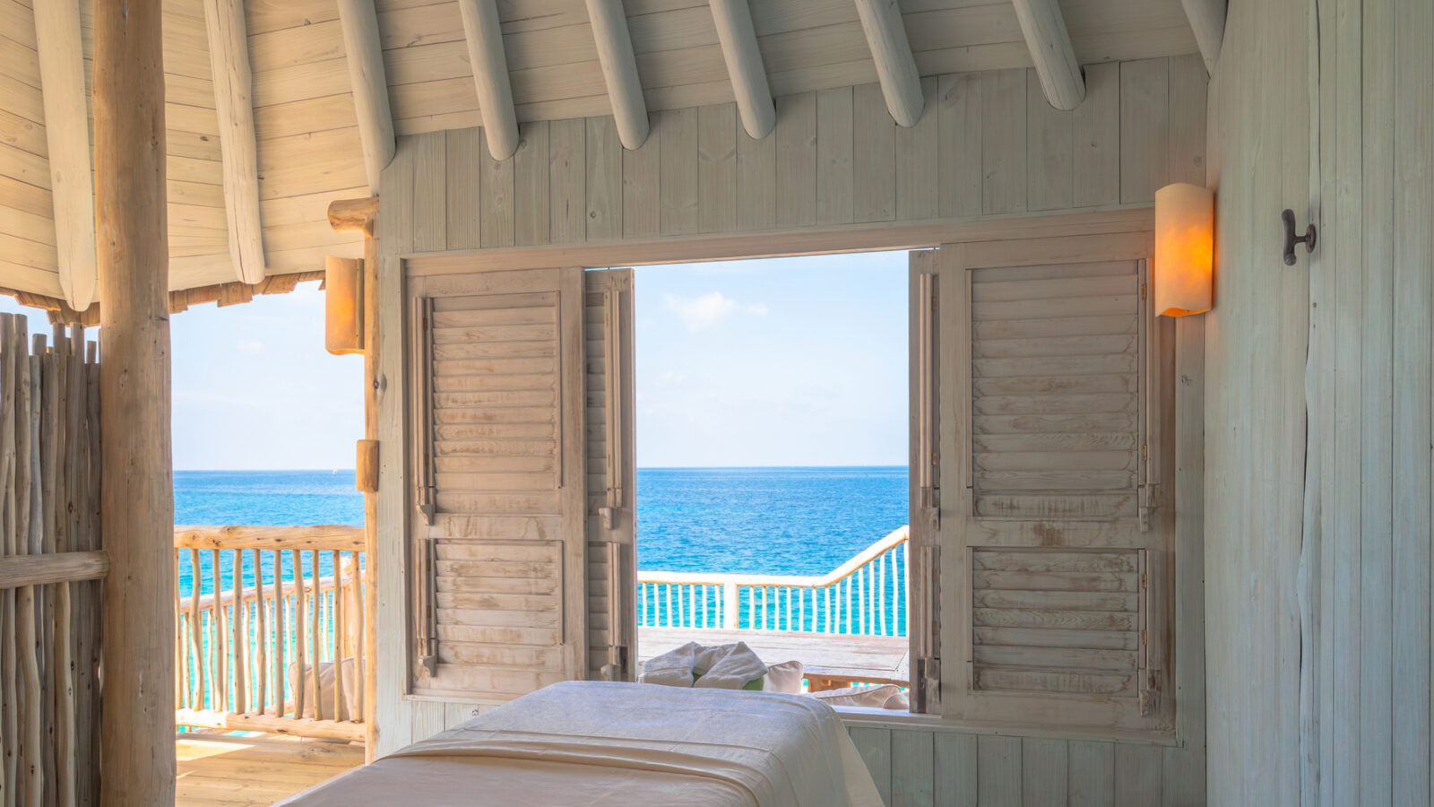 A serene beachside room at Crusoe Reserve features a massage table facing open wooden shutters, revealing a clear blue ocean view. The space is airy with white-washed wooden walls and soft ambient light, offering an idyllic escape.