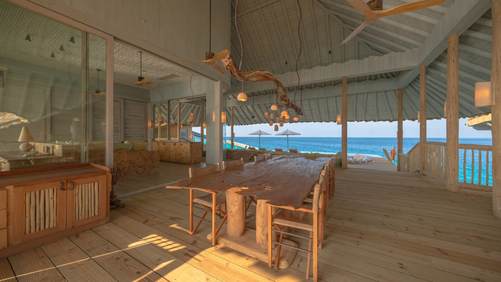 Spacious open-air wooden dining area at Crusoe Reserve overlooking the ocean. Features a long wooden table with chairs, a wooden ceiling with natural log supports, pendant lights, and ceiling fans. Glass panels separate it from an interior room, creating a bright, sunny atmosphere.