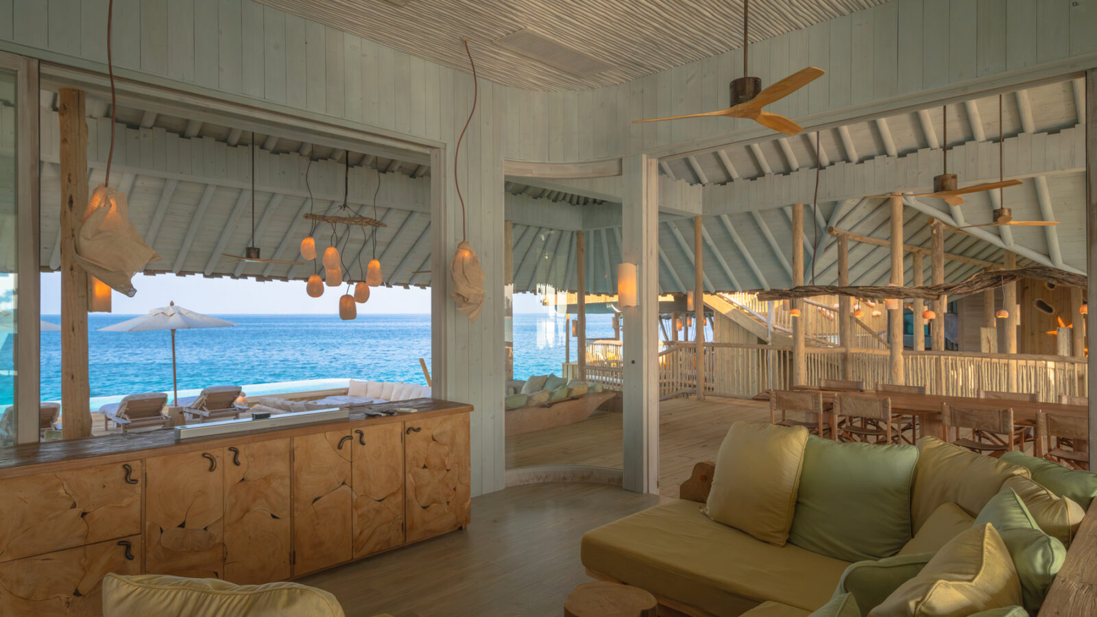 Crusoe Reserve’s tropical beachside lounge boasts open wooden architecture, green cushioned seating, and wooden tables. Through large windows, the blue ocean beckons. Hanging lanterns and a ceiling fan add charm, while a patio area with lounge chairs invites you to unwind outside.