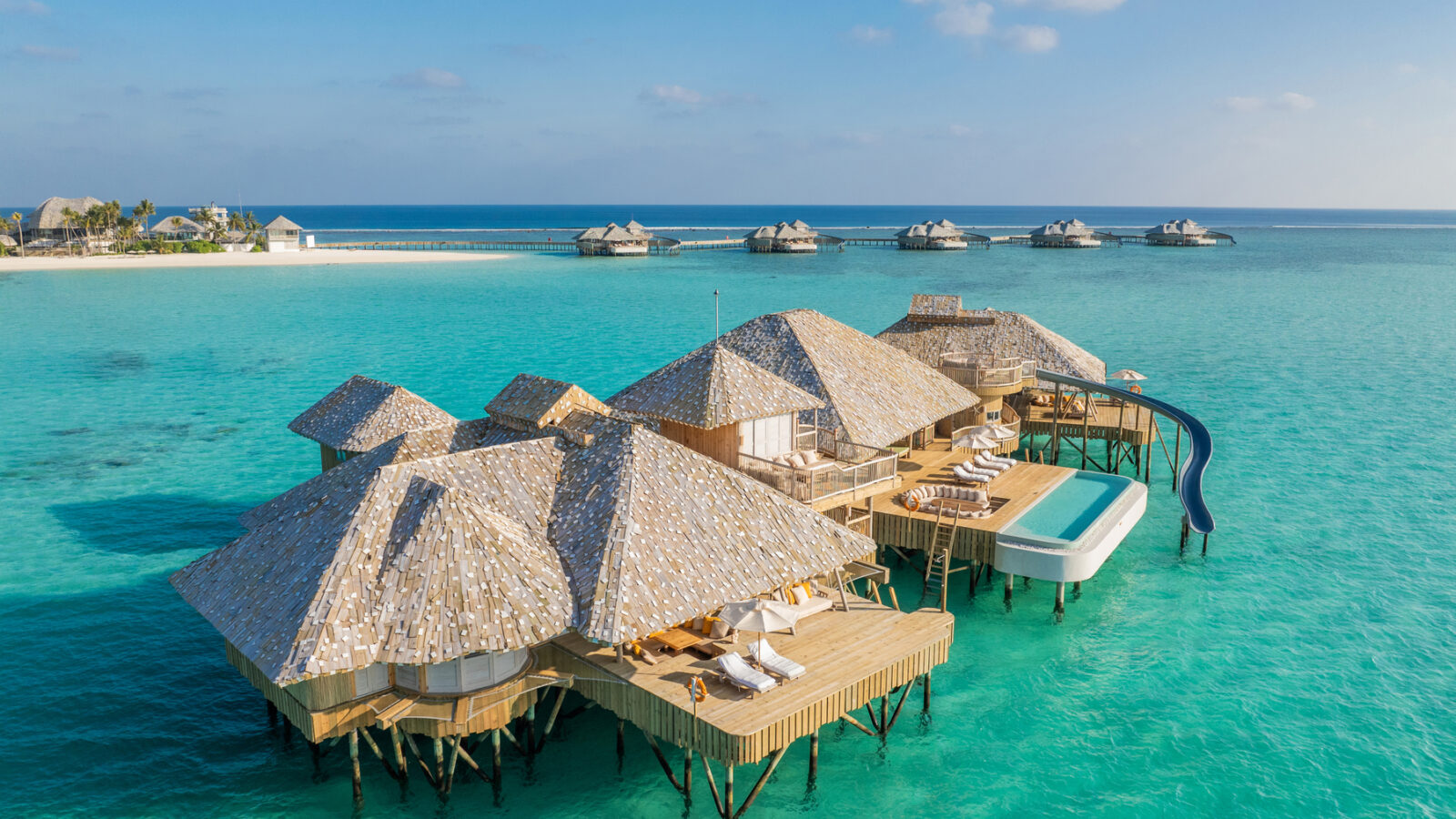 Aerial view of overwater bungalows with thatched roofs on a turquoise ocean. Each Crusoe Reserve bungalow features a private deck with lounge chairs and a slide leading into the water. White sand and more bungalows are visible in the distance under a clear blue sky.