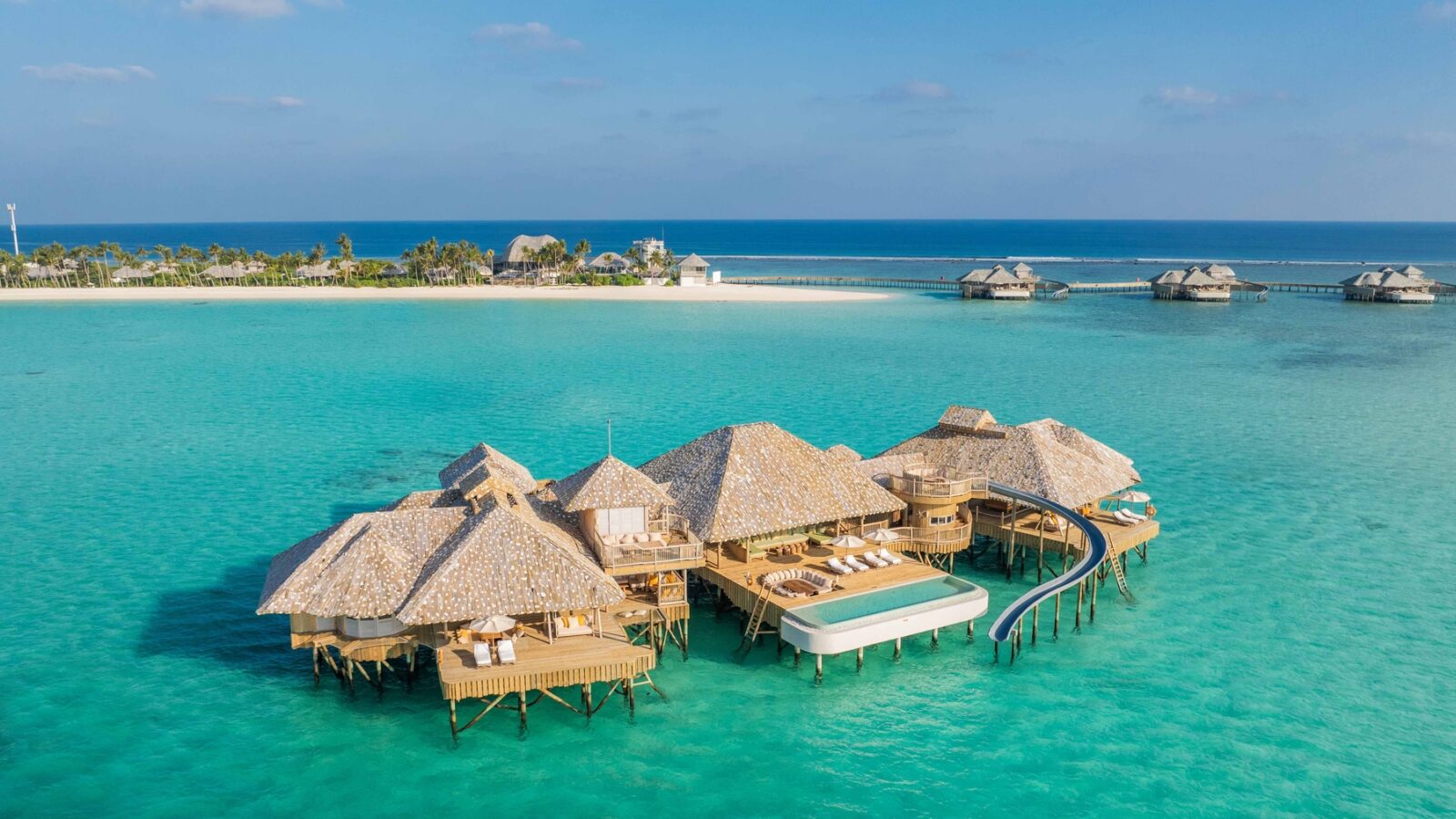 Aerial view of the Crusoe Reserve's luxurious overwater bungalows with thatched roofs in turquoise ocean waters. A curved slide extends from a deck into the sea. Sandy beaches and tropical greenery are visible in the background under a clear blue sky.