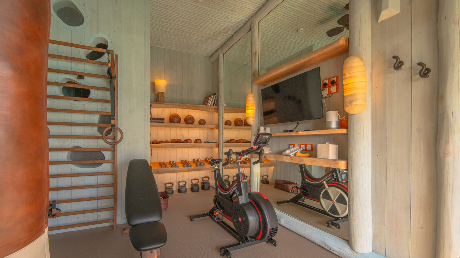 The home gym, akin to a Crusoe Reserve of fitness, features a punching bag, bench, stationary bike, and wall-mounted mirror. Shelves neatly hold free weights and other equipment. A ceiling fan and wall sconces create a warm and inviting atmosphere.