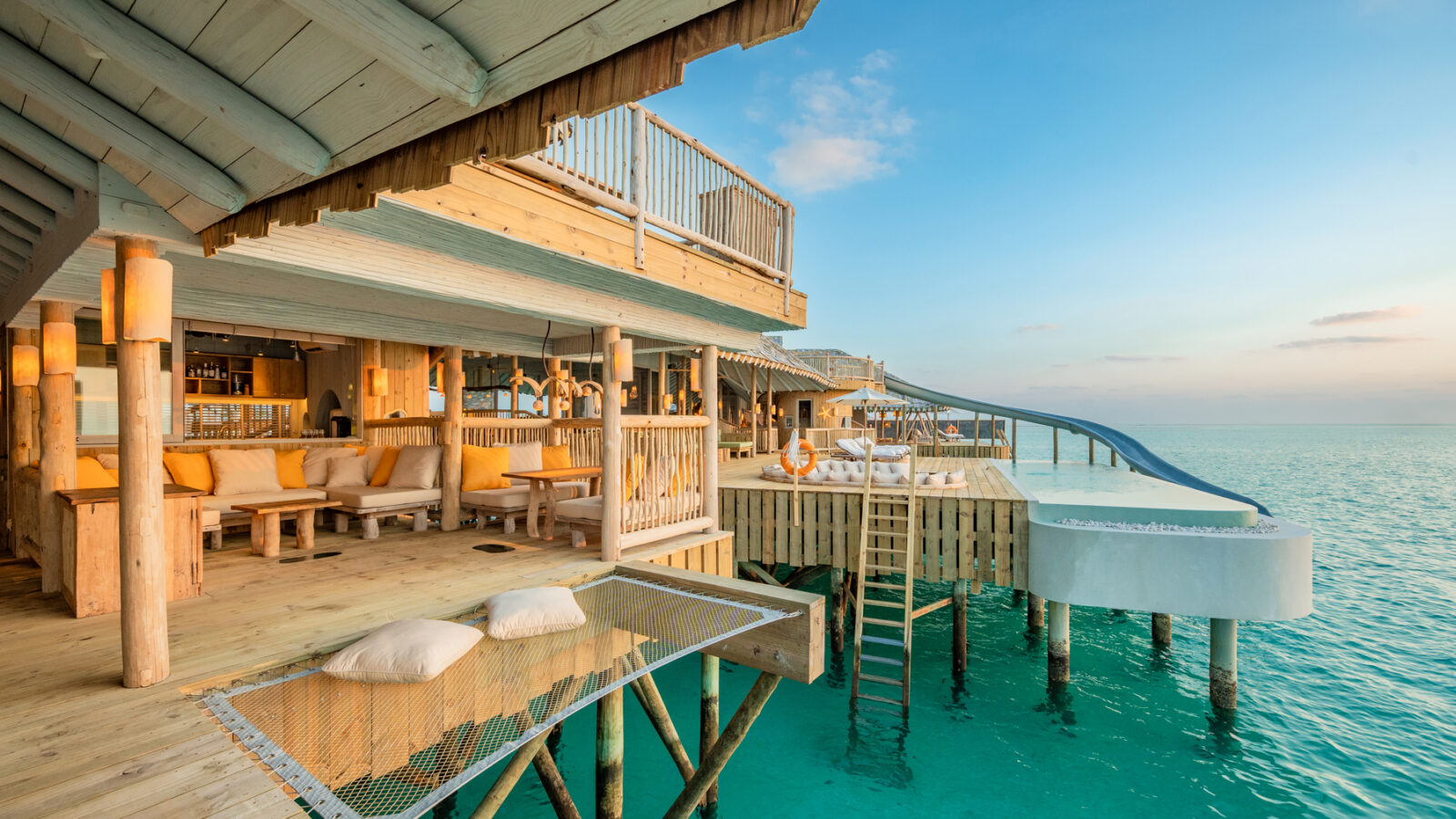 A luxurious Crusoe Reserve overwater villa features a wooden deck, hammock netting, and a slide into the clear blue ocean. The setting sun casts a warm glow over outdoor seating and dining areas, creating an idyllic escape.