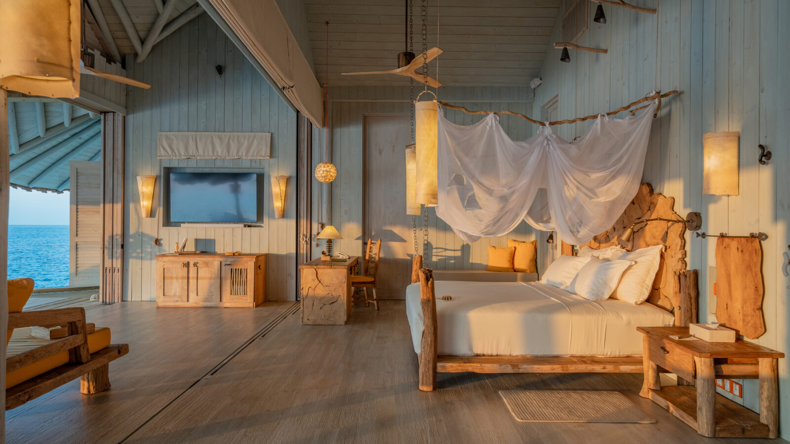 The Crusoe Reserve overwater bungalow offers a luxurious interior with rustic wooden furniture, a canopy bed adorned with white linens, and ambient lighting. Through open windows, enjoy the ocean view in a warm and inviting atmosphere.