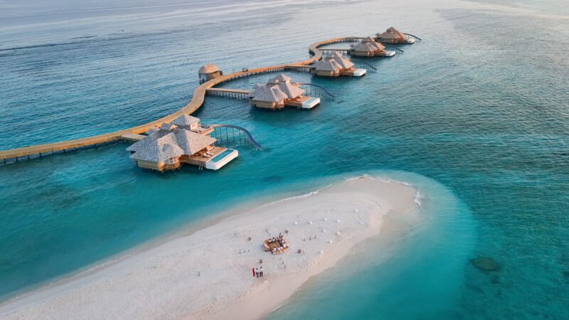 Aerial view of a Maldives luxury private resort with connected huts stretching out over a calm, turquoise sea. A small sandy island nearby hosts a group of people gathered around a picnic or event setup. The water is crystal clear, transitioning to deeper blue tones further out.