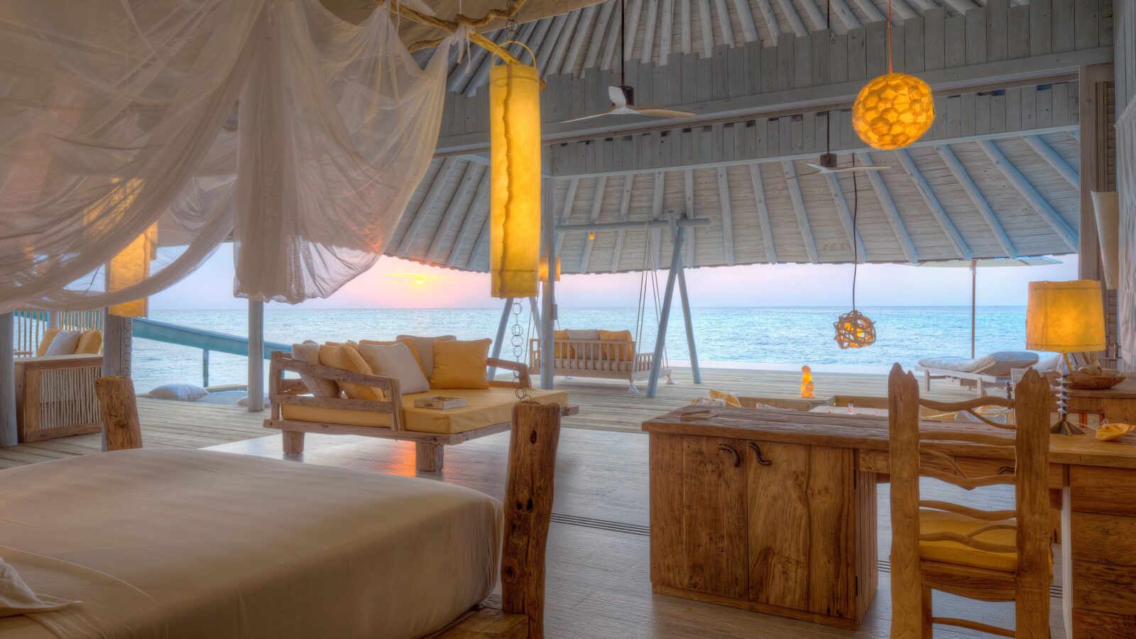 A luxurious oceanfront villa interior, a perfect beach hideaway with wooden furniture and warm lighting. A bed with a canopy sits on the left, while large windows reveal a serene sea view at sunset. Cozy seating areas and natural decor create a relaxing atmosphere.