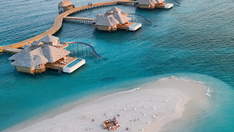 Aerial view of luxurious overwater bungalows connected by wooden walkways extending into the turquoise ocean at a Maldives private resort. In the foreground, a small sandy beach hosts a group with a picnic setup, surrounded by sun umbrellas. The serene sky is subtly lit with warm hues.