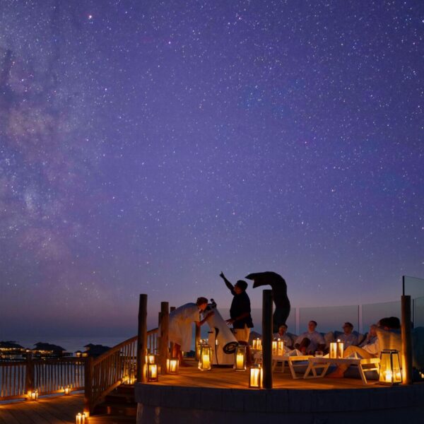 A group of people gather on a wooden deck at the Maldives' luxury private resort, Soneva, under a starry night sky. Candle lanterns illuminate the scene. A person on the deck points towards the sky, while others sit around a table, enjoying the view. The Milky Way is clearly visible in the background.