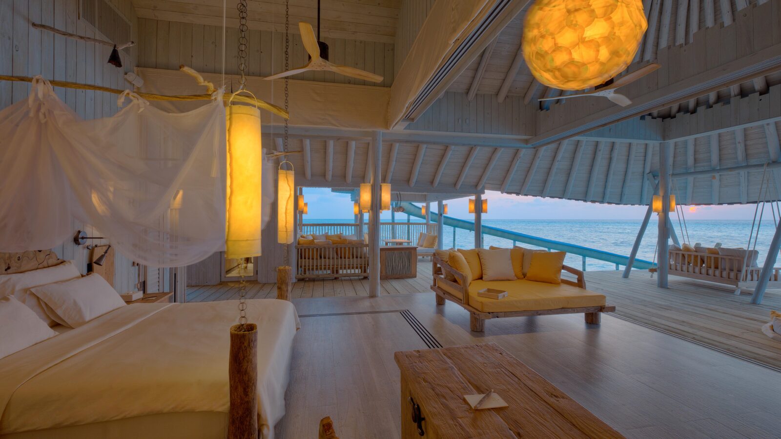 Discover the Soneva Secret 2024 overwater hideaway—a spacious, open-plan beach villa with a cozy bed draped in white netting, complemented by wooden furniture and soft yellow lighting. Large windows offer a stunning ocean view, creating a serene and inviting atmosphere.