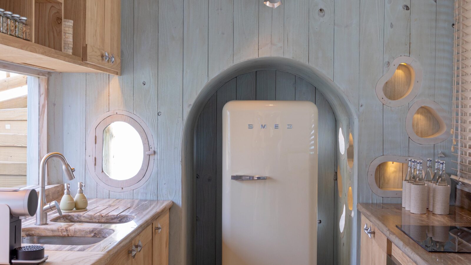 A cozy, small kitchen with light wood cabinets and walls, reminiscent of a Soneva Secret 2024 overwater hideaway. It features a vintage-style refrigerator, a round window, and modern lighting. Dishes and appliances are neatly arranged on countertops and shelves.