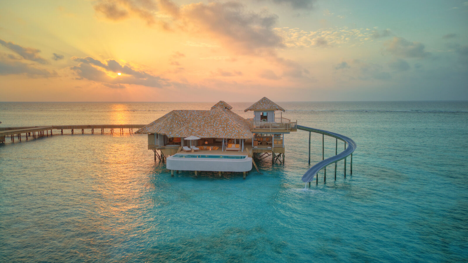 Discover the Soneva Secret 2024 overwater hideaway, a luxurious villa in a tropical paradise at sunset. With thatched roofs, a private pool, and a waterslide leading into the clear blue ocean, it's pure bliss. A wooden walkway connects it to shore under vibrant orange and pink skies.