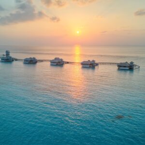 A serene view of a sunset over the ocean at a Maldives luxury private resort, featuring a row of thatched-roof overwater bungalows connected by a wooden pier. The sun is low on the horizon, casting a warm orange glow on the calm waters. The sky is lightly clouded, enhancing the peaceful scene.