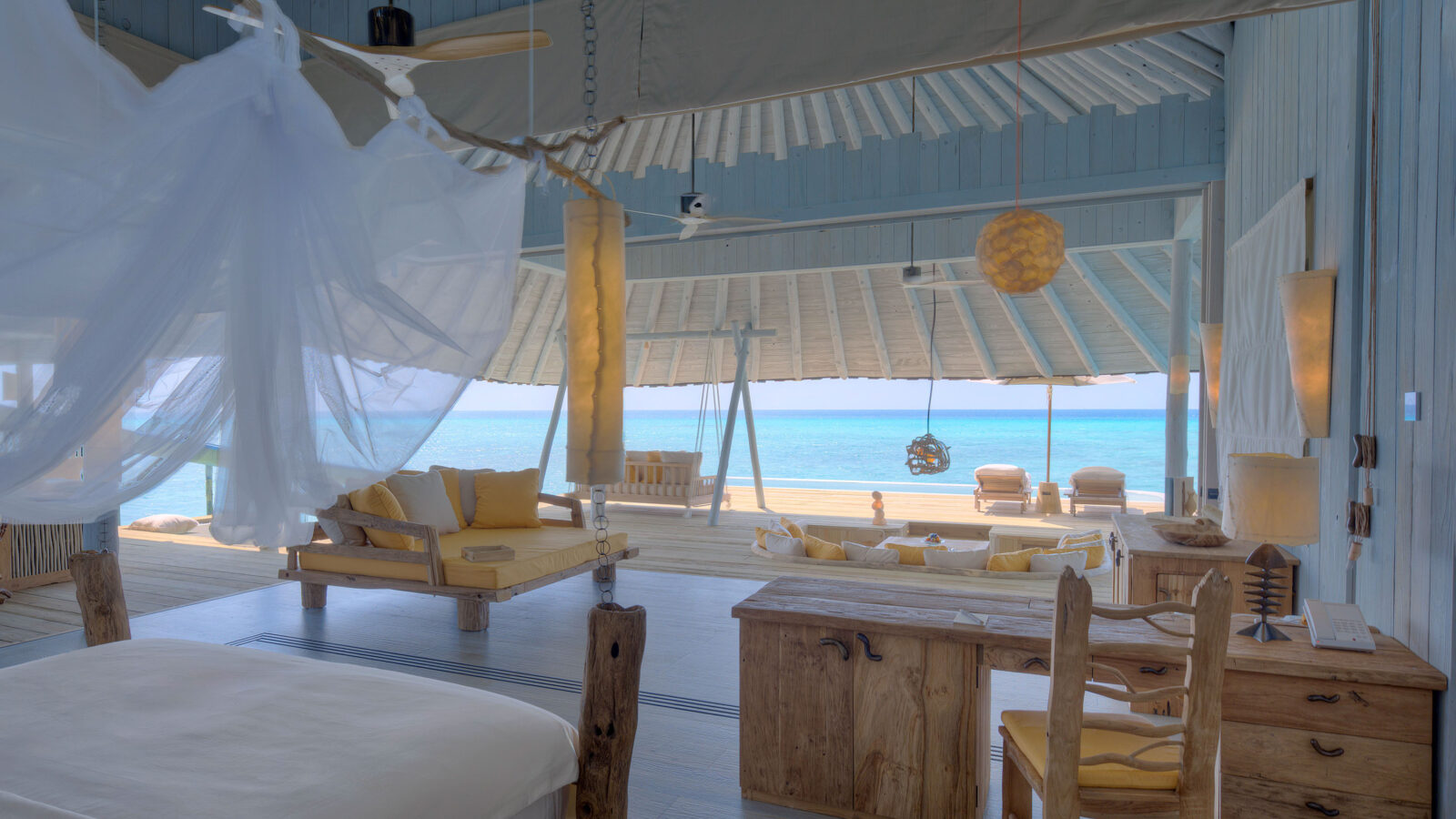 A spacious, open-air beachfront bedroom with rustic wooden furniture, white bedding, and a canopy net. The room opens out to a stunning ocean view, with cozy seating areas, a wooden swing chair, and natural light flooding in from large windows and doors. Enjoy Soneva Exclusive Offers for the ultimate luxury stay.