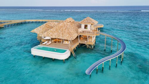 A luxurious overwater villa at the Maldives' private resort, Soneva, stands on stilts in a clear, turquoise sea. It features thatched roofs, a private infinity pool, and a long slide leading directly into the water. Wooden walkways connect the villa to other similar structures in the distance.