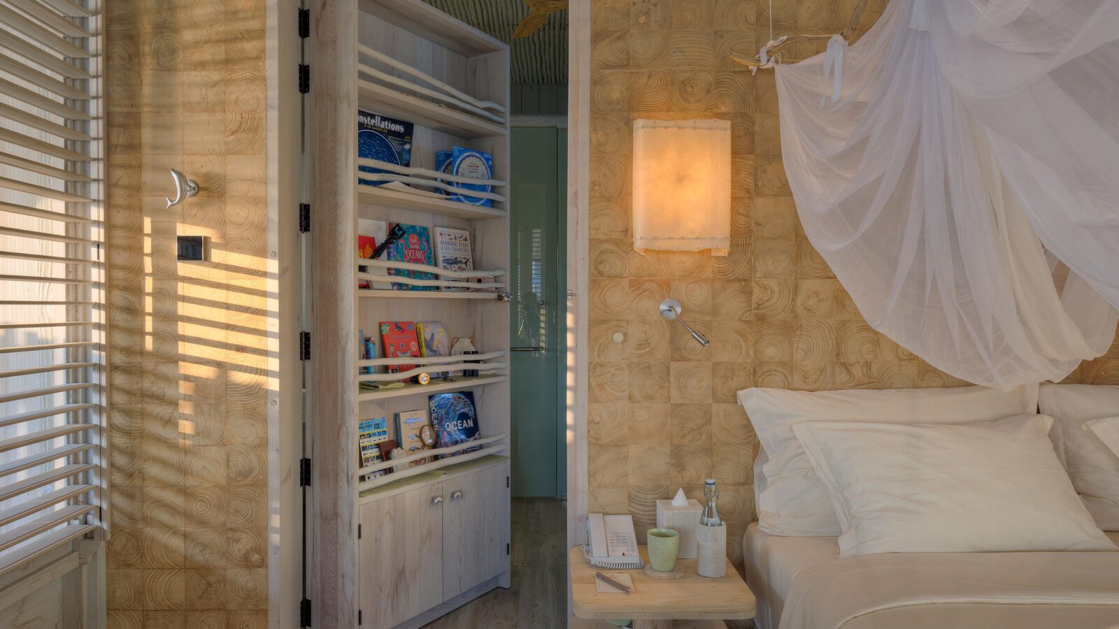 A cozy bedroom with beige and light wood decor has a bed with white bedding and a mosquito net. To the left, an open door reveals a small bookshelf filled with books. A nightstand next to the bed holds water bottles, a glass, and a book. Experience tranquility redefined with Soneva Exclusive Offers. Light filters through blinds.