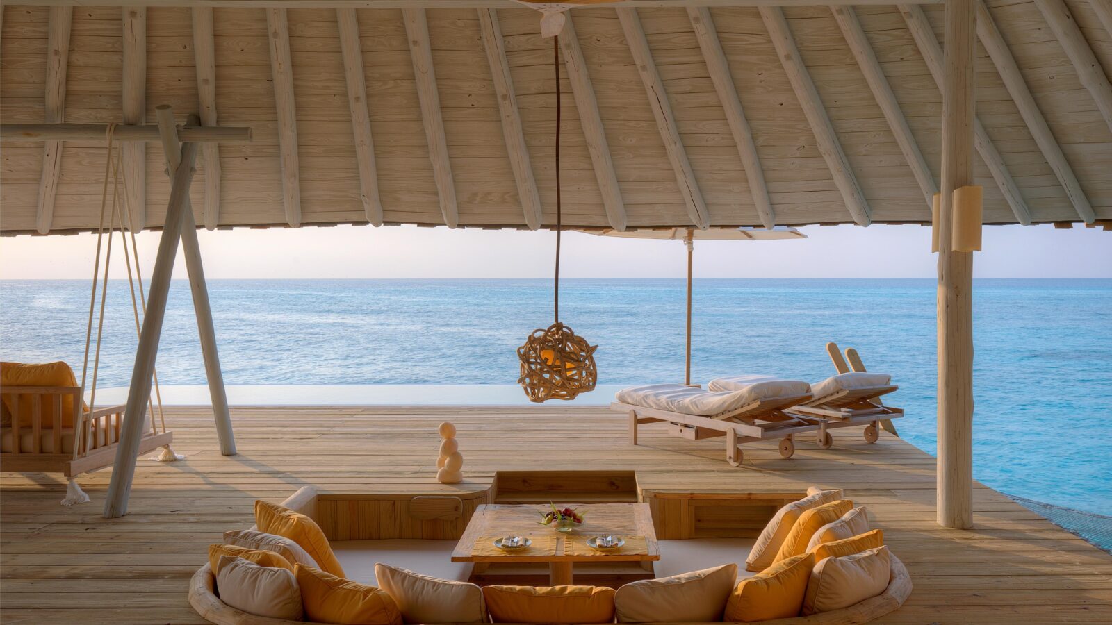 Wooden deck overlooking a vast, calm ocean. The deck features a sunken seating area with yellow cushions around a low table, a hanging woven light fixture, and two lounge chairs facing the water. A slanted roof provides shade, and an overhead ceiling fan adds a breezy touch—a slice of paradise inspired by Soneva Exclusive Offers.