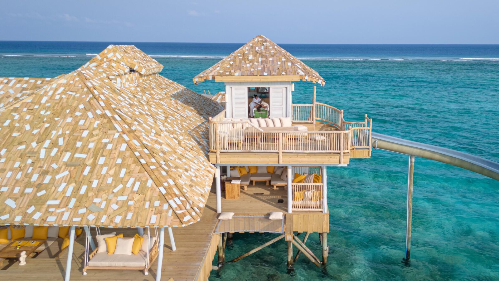 An overwater bungalow with thatched roofs at a Maldives luxury private resort features multiple outdoor seating areas, a slide leading directly into the vibrant blue ocean, and a person standing on the upper deck enjoying the view. The scene is surrounded by clear turquoise waters under a sunny sky.