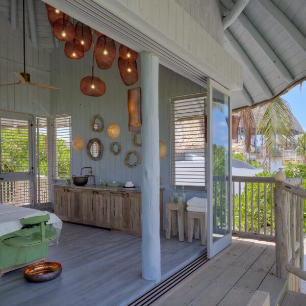 A serene spa room with open shutters overlooks lush greenery and palm trees. The room features a massage table with a white sheet and green blanket, wooden furniture, hanging wicker light fixtures, and a basin with a bronze bowl on the floor. Natural light brightens the space, perfect for Soneva Exclusive Offers.